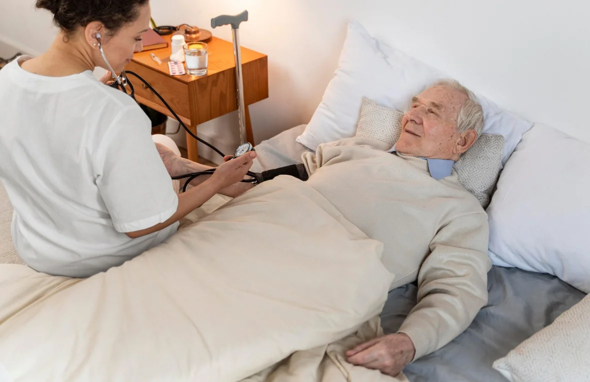 A caregiver checks the blood pressure of an elderly man who is lying in bed, with medical supplies placed nearby on a bedside table.