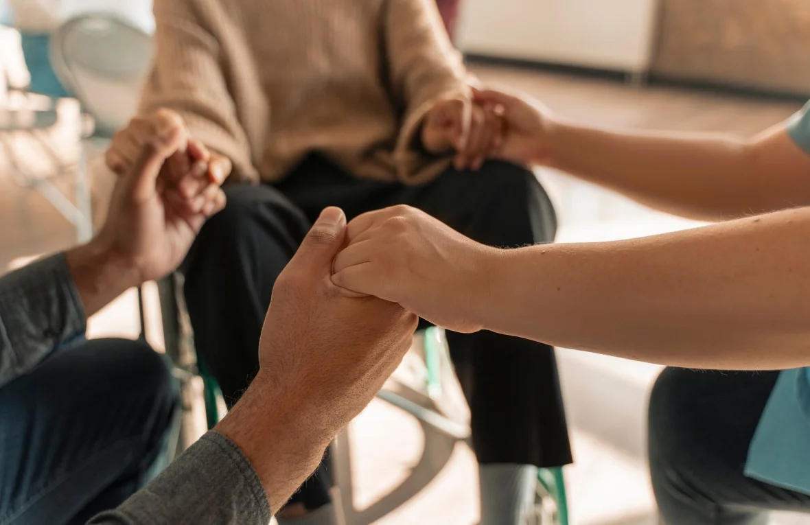 Several people hold hands in a comforting gesture, providing emotional support, likely in a care or therapy setting.