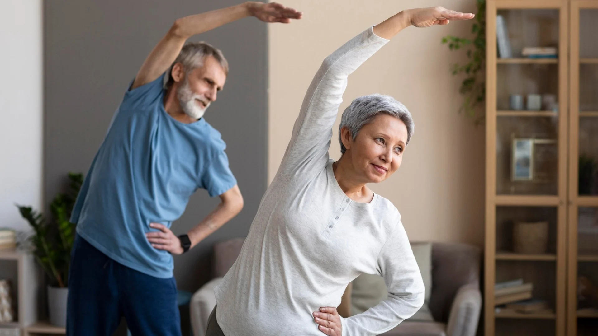 Elderly woman and man stretching arms together at home.