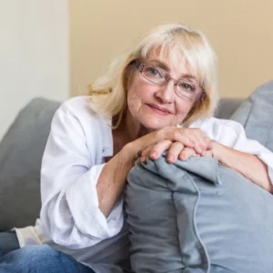 An elderly woman in a white shirt and cozy knit vest sits on a couch, leaning forward and holding a pillow with her hands resting on it.
