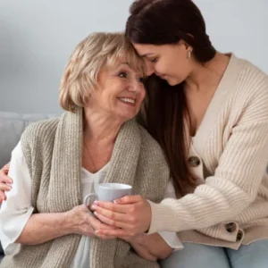 An elderly woman holding a cup sits closely with a younger woman in a cream sweater, who lovingly embraces her and rests her head affectionately on the elder's shoulder.
