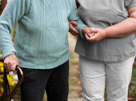 A caregiver provides physical support to an elderly person using a walking stick, holding the individual's arm and hand while walking together.