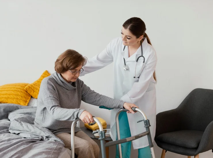 A doctor in a white coat helps an elderly woman using a walker move from her bed, offering support and encouragement.