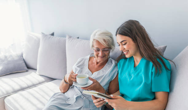 Happy patient is holding caregiver for a hand while spending time together. Elderly woman in nursing home and nurse. Aged elegant woman and tea time at nursing home