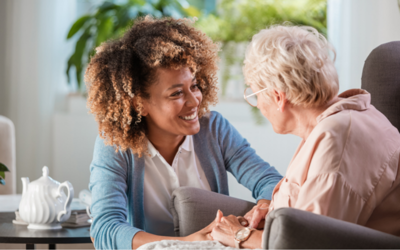 A caregiver offers emotional support and holds hands with an elderly woman, having a comforting conversation in a bright living room.