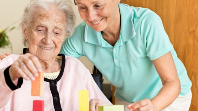 A caregiver assists an elderly woman with therapeutic activities, guiding her as she stacks colorful blocks on a table.