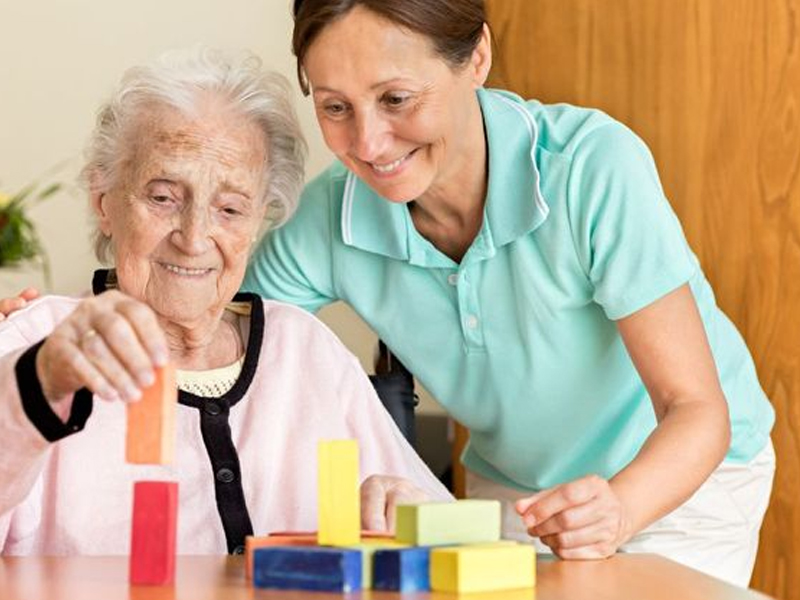 A caregiver assists an elderly woman with therapeutic activities, guiding her as she stacks colorful blocks on a table.