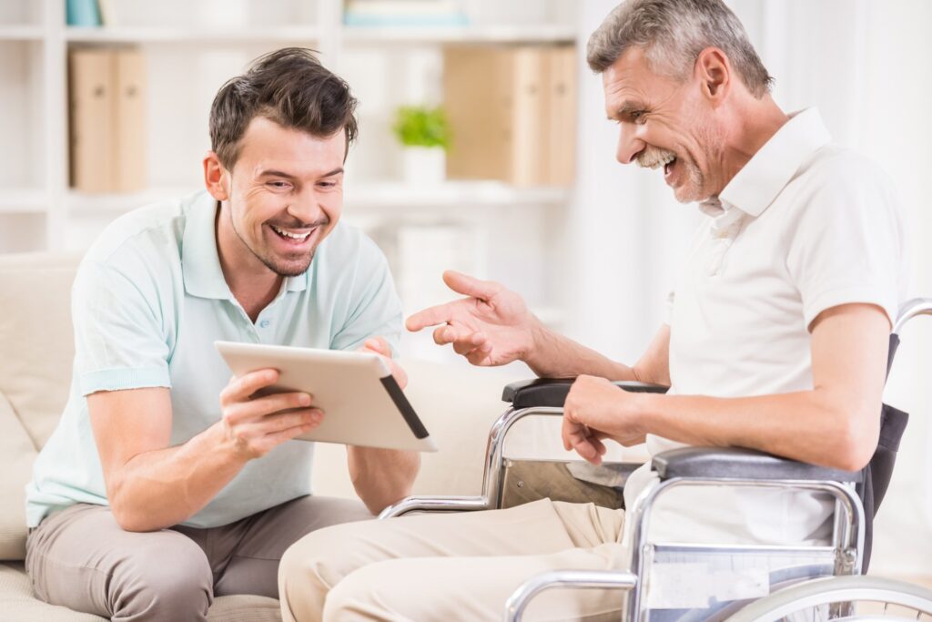A caregiver sits and discusses something on a tablet with an elderly man in a wheelchair in a bright room.