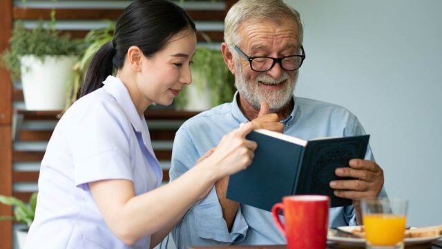 A caregiver discuss a book with an elderly man over a cup of tea in a comfortable indoor setting.