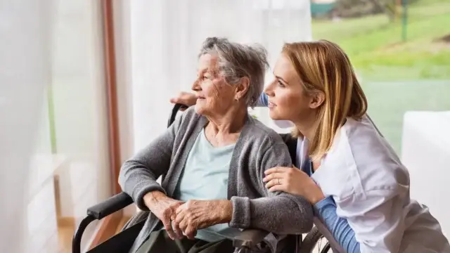 A caregiver in a white coat and an elderly woman in a wheelchair enjoy a peaceful moment together, looking out the window.