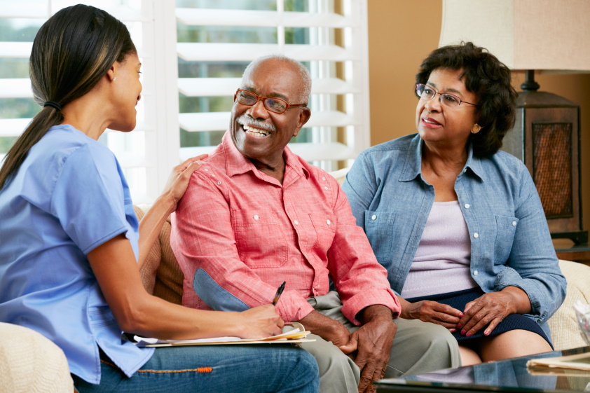 A nurse in blue scrubs talks with an elderly man and woman, both smiling, during a meeting in a comfortable living room.