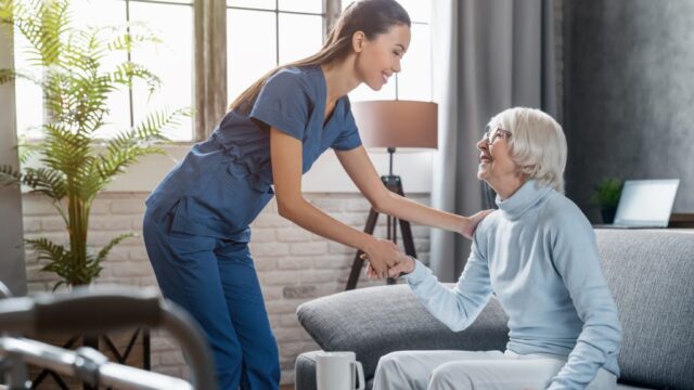 A caregiver in blue scrubs smiles while helping an elderly woman in a light blue sweater, both holding hands in a warm moment.