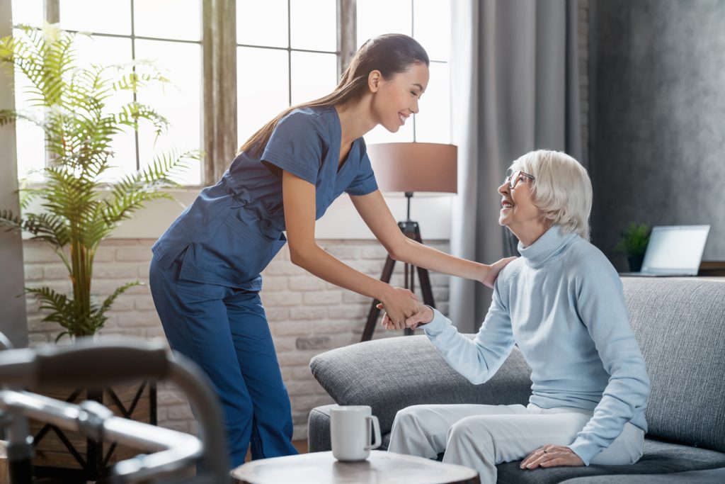 A caregiver in blue scrubs smiles while helping an elderly woman in a light blue sweater, both holding hands in a warm moment.