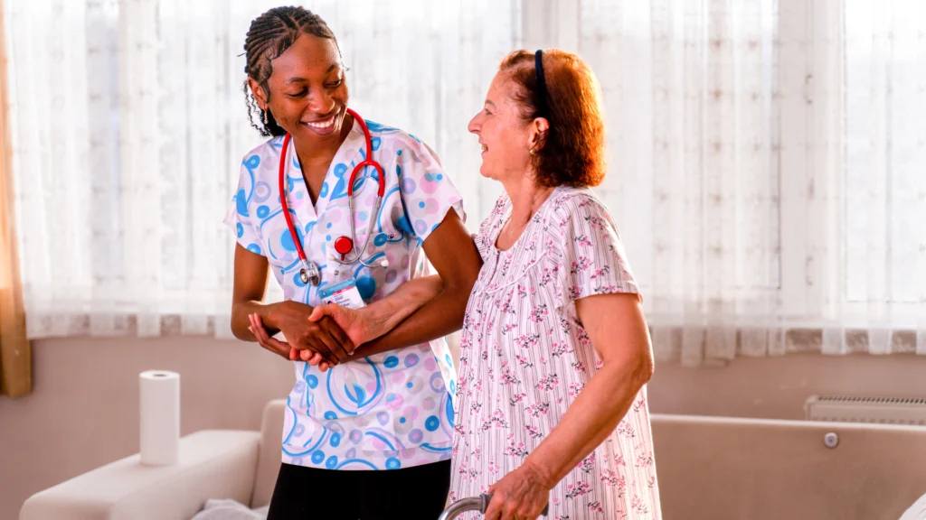 A caregiver in a colorful outfit assists an elderly woman in a dress, both smiling and walking together.