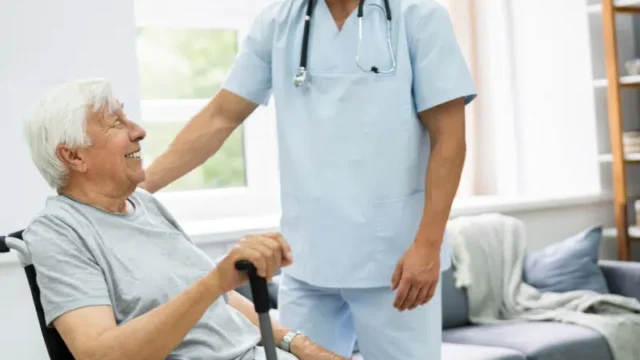 A healthcare professional in a blue uniform assists an elderly man in a wheelchair, both sharing a friendly moment.