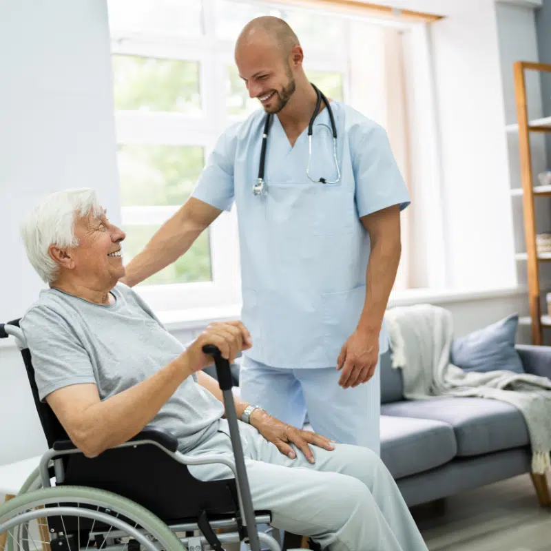 A healthcare professional in a blue uniform assists an elderly man in a wheelchair, both sharing a friendly moment.