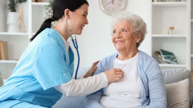 A caregiver in blue scrubs smiles at an elderly woman while adjusting a stethoscope on her chest.
