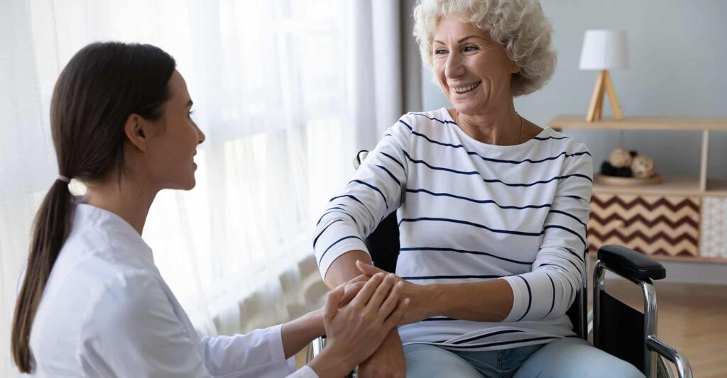 A caregiver in white scrubs holds hands with an elderly woman in a wheelchair, sharing a joyful moment together.
