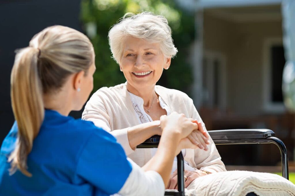 Nurse chatting with elderly woman in wheelchair outdoors.