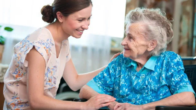 A caregiver smiles at an elderly woman in a wheelchair, sharing a warm and caring moment together.
