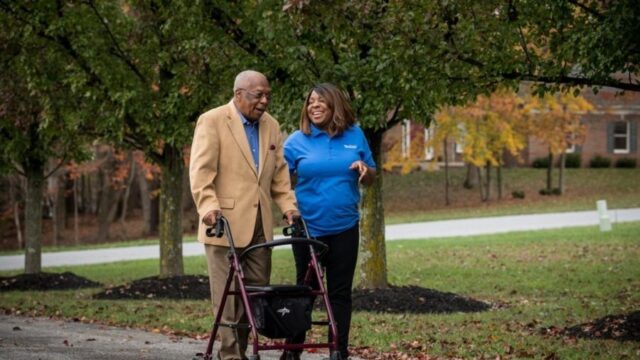 Elderly man using walker with companion in park.