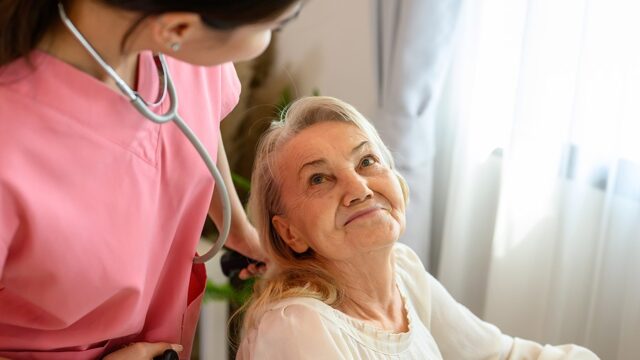 A caregiver in pink scrubs adjusts the stethoscope as she smiles at an elderly woman in a wheelchair.