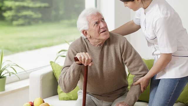 A caregiver assists an elderly man with a cane, offering support as he looks up at her.