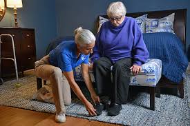 A caregiver kneels and helps an elderly woman put on her shoes while she sits on the bed.