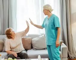 A nurse in blue scrubs and an elderly woman sitting on a couch give each other a high-five in a living room.