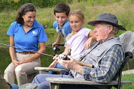 An elderly man sits fishing by a pond with a group of people, including a caregiver in a blue shirt.