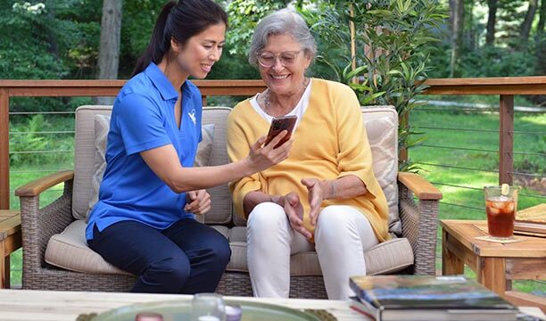 A caregiver in a blue shirt shows an elderly woman something on a phone, both smiling and enjoying a moment together outdoors.
