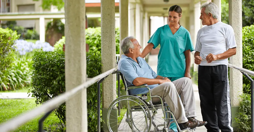 A caregiver in teal scrubs chats with two elderly men, one in a wheelchair and the other standing, in a pleasant outdoor setting.