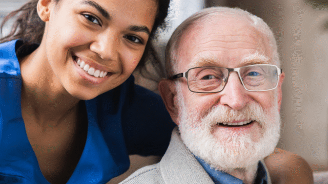 A caregiver and an elderly man smile together, sharing a heartwarming moment on a couch.