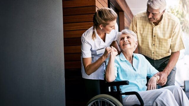 A caregiver in a white shirt helps an elderly woman in a wheelchair while a man stands beside her, both offering support.