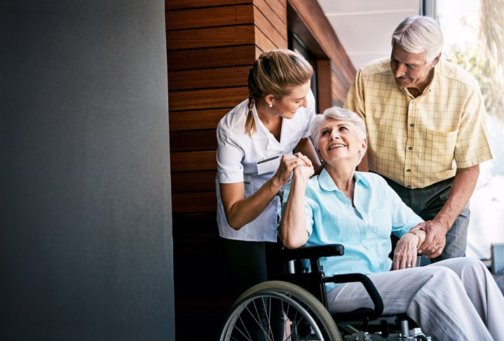 A caregiver in a white shirt helps an elderly woman in a wheelchair while a man stands beside her, both offering support.