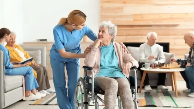 A caregiver in blue scrubs gently assists an elderly woman in a wheelchair while other seniors relax in the background.