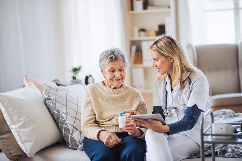 A caregiver in a white uniform shows medication instructions on a tablet to an elderly woman holding a pill bottle.