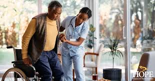 A nurse helps an elderly man stand up from his wheelchair in a bright, well-lit room.