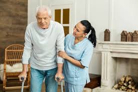 A caregiver helps an elderly man using crutches stand in a living room.