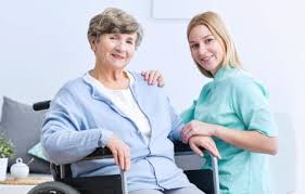 A nurse in green uniform supports and talks with an elderly woman in a wheelchair in a bright room.