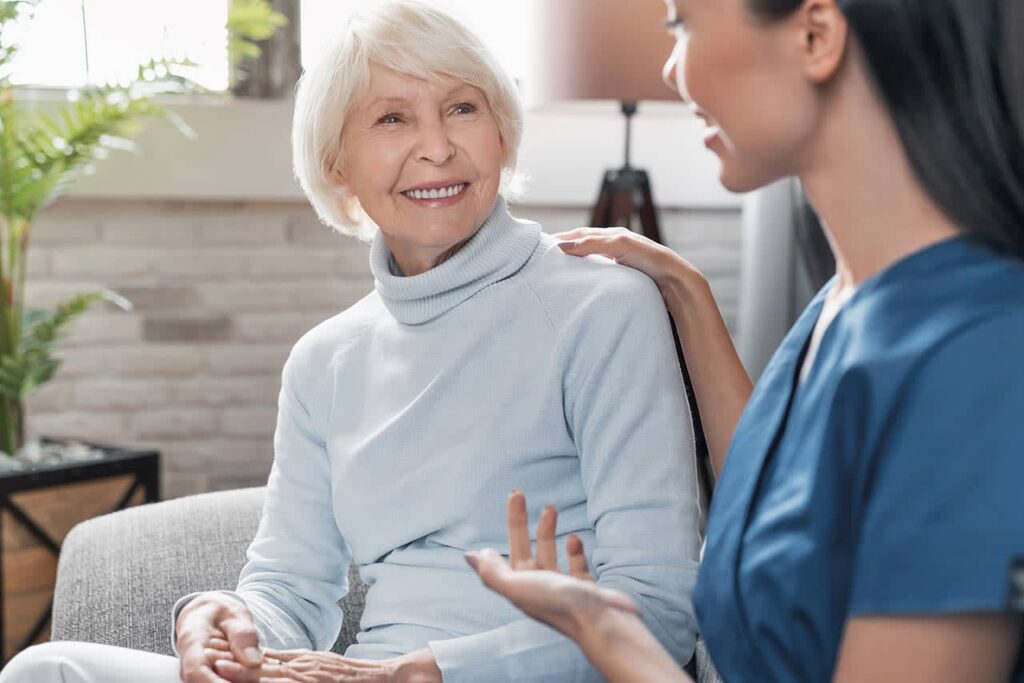A caregiver offers support to an elderly woman in a cozy living room, helping her with her daily needs.