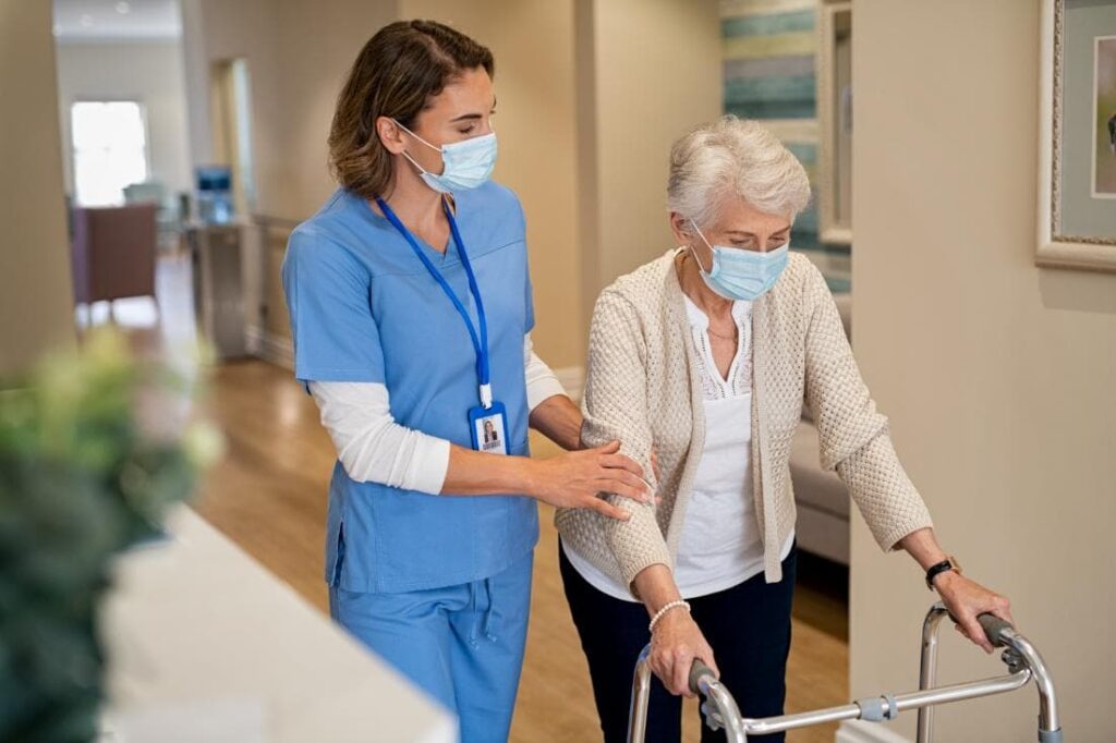 A caregiver in blue scrubs assists an elderly woman with a walker, both wearing masks while walking in a hallway.